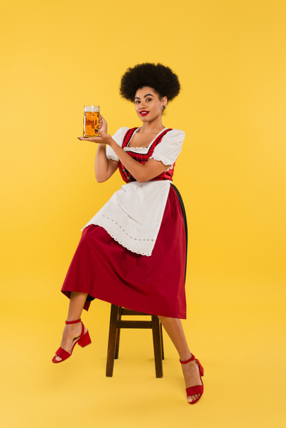 молода афроамериканська офіціантка в автентичній баварській сукні, що сидить на стільці з пивним кухольком на жовтому - Фото, зображення