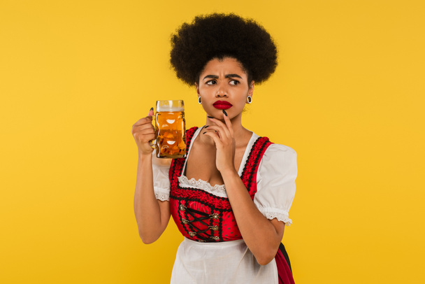 pensativo africano americano oktoberfest camarera en dirndl de pie con taza de cerveza en amarillo - Foto, Imagen