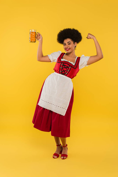 vzrušený africký americký oktoberfest servírka s pivem hrnek ukazující vítězství gesto na žluté - Fotografie, Obrázek