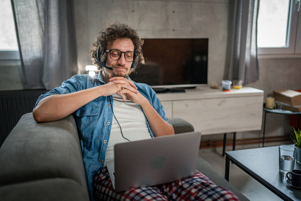Un homme caucasien travail indépendant masculin de la maison avec des écouteurs sur sa tête sur ordinateur portable répartiteur de soutien à la clientèle tandis que s'asseoir sur le canapé-lit à la maison en chemise et pyjama sourire heureux confiant - Photo, image