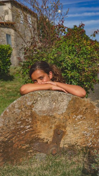 girl with her arms resting on a millstone in a garden with a stone construction in the background - Photo, Image