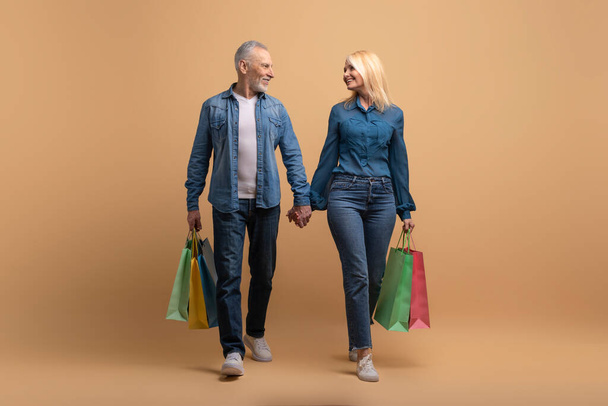 Όμορφη ευτυχισμένη συνταξιούχος ζευγάρι άνδρας και γυναίκα σε casual περπάτημα από το εμπορικό κέντρο, κρατώντας τα χέρια, κοιτάζοντας ο ένας τον άλλον και χαμογελώντας, μεταφέρουν πολύχρωμες αγορές, μπεζ φόντο, πλήρες μήκος - Φωτογραφία, εικόνα