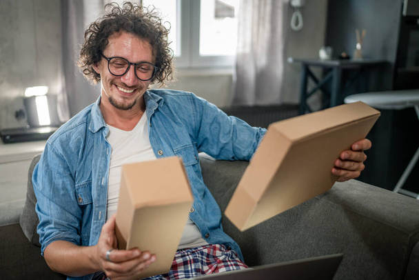 jeden biały mężczyzna dorosły mężczyzna siedzieć w domu na kanapie otrzymując pakiet pocztą produkt zamówił online lub specjalny prezent szczęśliwy uśmiech prawdziwy ludzie dostawy koncepcji usługi - Zdjęcie, obraz