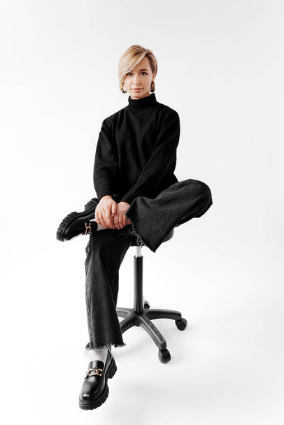 Повне тіло молодої стильної блондинки сидить на стільці і дивиться на камеру. Вона одягнена в стильний і модний светр чорних черепах, чорні штани та чорне взуття на білому тлі - Фото, зображення