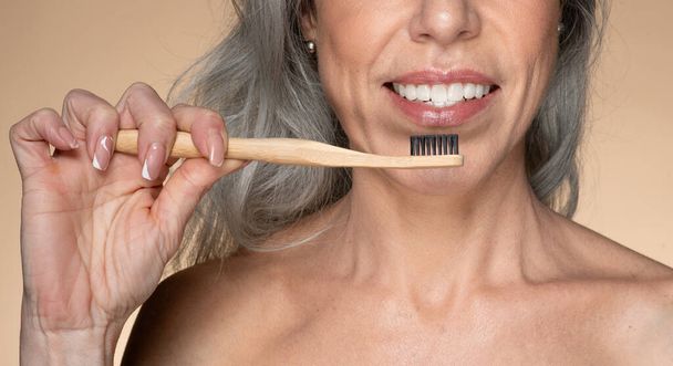 Mujer mayor feliz utilizando el cepillo de dientes de bambú natural ecológico para limpiar los dientes, disfrutando de artículos de plástico gratis para la higiene personal, de pie sobre fondo beige, primer plano - Foto, Imagen