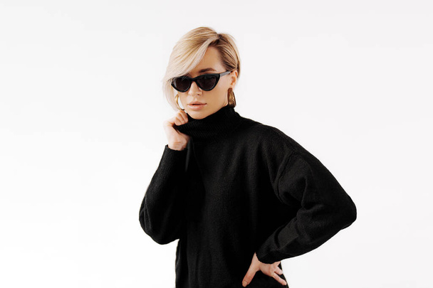 Una donna sicura ed elegante che indossa un maglione nero che contrasta con i suoi capelli biondi e lo sfondo bianco. Questa foto può essere utilizzata per vari scopi, come pubblicità, moda o stile di vita - Foto, immagini