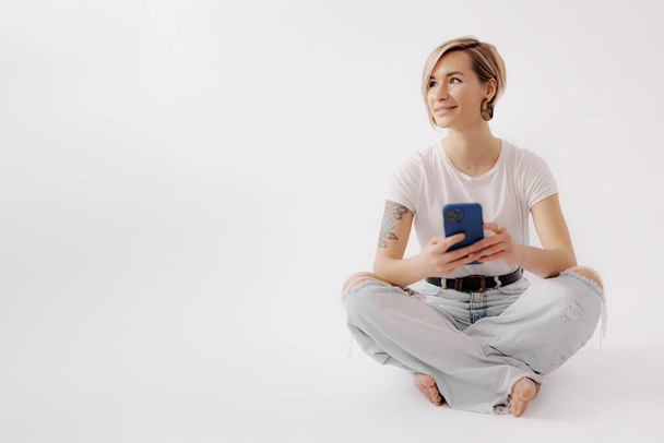 Νεαρή γυναίκα που κάθεται στο πάτωμα κρατώντας ένα smartphone στα χέρια της. Φοράει λευκό μπλουζάκι και σιέλ τζιν, και έχει τατουάζ στο δεξί της χέρι. Δείχνει χαλαρή και χαρούμενη. - Φωτογραφία, εικόνα