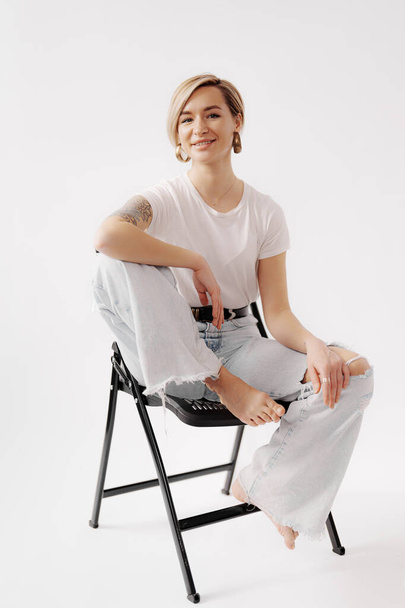 Усміхнена жінка сидить на чорному стільці з руками на колінах. На ній біла футболка, розірвані джинси. Вона має татуювання на руках і позує в розслабленому вигляді - Фото, зображення