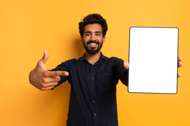 Oferta en línea. Hombre indio guapo apuntando a la tableta digital con pantalla blanca, sonriendo joven oriental que demuestra espacio libre de la copia para el diseño del anuncio, de pie sobre fondo amarillo, burla - Foto, Imagen