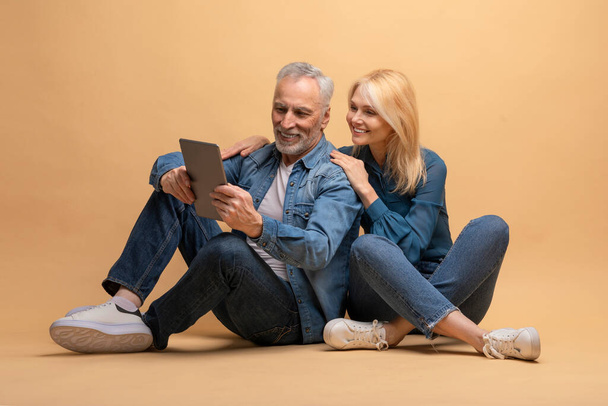 Positives älteres Paar, das auf dem Boden sitzt, sich umarmt und ein modernes digitales Tablet nutzt, isoliert auf beigem Studiohintergrund. Mann und Frau im Ruhestand surfen, scrollen, online einkaufen auf Pad - Foto, Bild