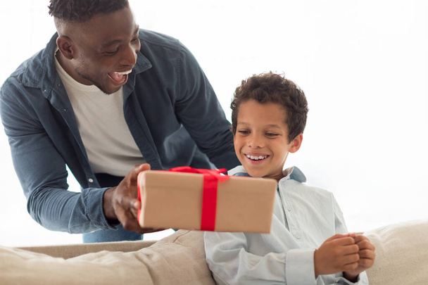 Liebevoller afrikanisch-amerikanischer Papa schenkt seinem kleinen Sohn eine Geschenkbox, grüßt ihn und gratuliert ihm zum Geburtstag im modernen Heim. Familie feiert Urlaub, Vater beschenkt Kind - Foto, Bild
