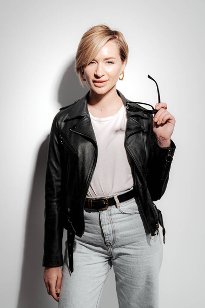Mujer joven con estilo y confianza posando frente a una pared blanca con una chaqueta de cuero negro. Ella sostiene un par de gafas de sol en una mano y mira a la cámara con una expresión segura - Foto, Imagen