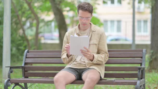 Jeune homme occasionnel lisant des documents assis à l'extérieur - Séquence, vidéo