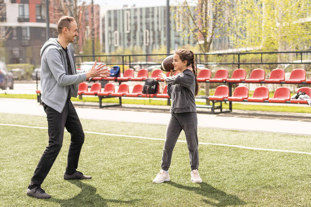 Mädchen hält einen ovalen braunen Leder-Rugby-Ball in der Hand und lächelt, während sie mit ihren Eltern im Park spielt. Familie und Kinder, Naturkonzept. - Foto, Bild