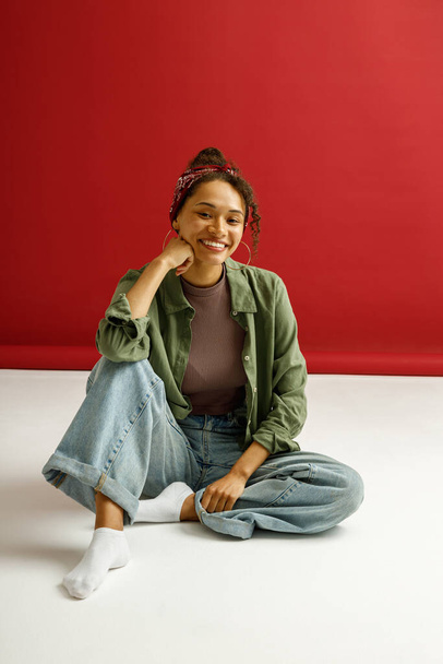 Nainen istuu lattialla ja näyttää kameralta hymy punaisen studion taustalla. Laadukas kuva - Valokuva, kuva