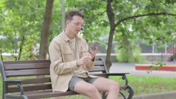 Современный молодой человек разговаривает по телефону, сидя на скамейке - Кадры, видео