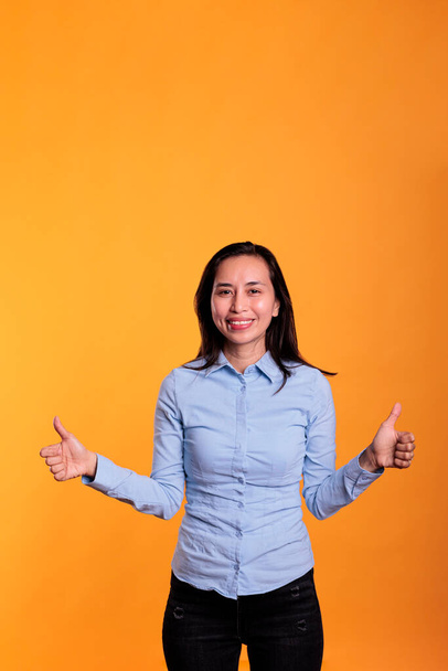 Mujer asiática sonriente haciendo un gesto de pulgares hacia arriba delante de la cámara mostrando aprobación y gesto de acuerdo con los dedos en el estudio. Modelo satisfecho gesto bien y símbolo positivo sobre fondo amarillo - Foto, Imagen