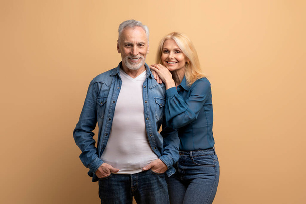Liebe, Ehe, Zuneigung, Beziehungskonzept. Glücklich verheiratete ältere kaukasische Paar ältere Mann und Frau in Jeans-Outfits posieren umarmend und lächelnd in die Kamera auf beigem Hintergrund - Foto, Bild
