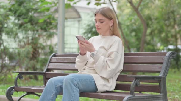 Casual giovane donna che celebra il successo online sul telefono mentre si siede all'aperto - Filmati, video