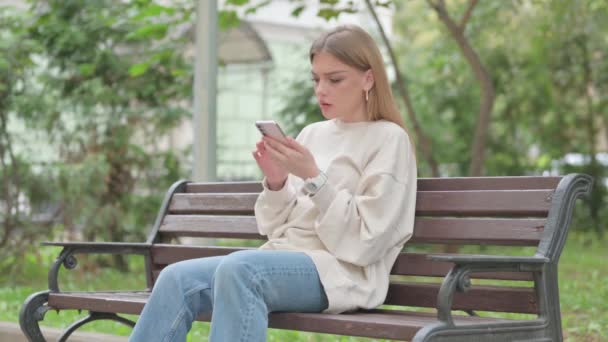 Casual jonge vrouw geschokt door verlies op telefoon tijdens het zitten buiten op een bank - Video