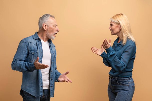 Unhappy marriage, crisis in relationships. Angry senior blonde woman wife have quarrel with her furious husband grey-haired man, elderly couple fighting over beige studio background - Foto, Imagen