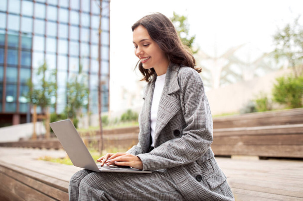 Фріланс, інтернет-бізнес. Усміхнена молода бізнес-леді, використовуючи ноутбук, працює і робить відеодзвінки через комп'ютер, сидячи в міській місцевості зовні. Вид збоку бізнес-леді, що друкує на ПК на відкритому повітрі - Фото, зображення
