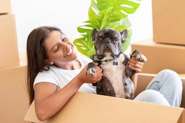 Closeup of pretty young woman with french bulldog in empty house among cardboxes with belongings, playing with her pet dog in new apartment, enjoying new house. Rent, relocation, moving - Photo, Image