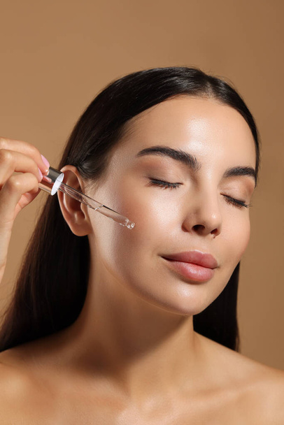 Beautiful young woman applying serum onto her face on beige background, closeup - Photo, Image