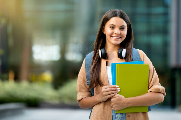 Pozytywna młoda studentka ubrana w luźny strój z plecakiem i notatnikami do ćwiczeń pozująca na świeżym powietrzu na kampusie uniwersyteckim, uśmiechnięta do kamery, przestrzeń do kopiowania. Styl życia studentów - Zdjęcie, obraz