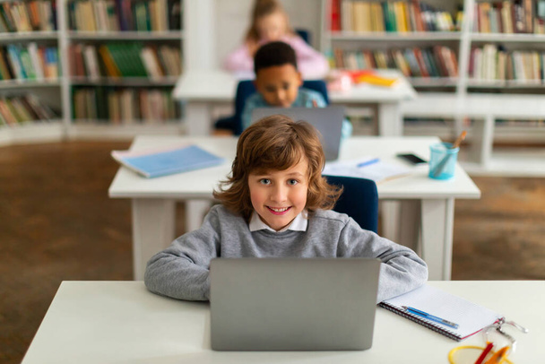Nowoczesna edukacja podstawowa. Szczęśliwe dzieci w szkole siedzi przy biurkach z laptopami, studiuje za pomocą cyfrowych gadżetów, chłopiec patrzy i uśmiecha się do kamery - Zdjęcie, obraz