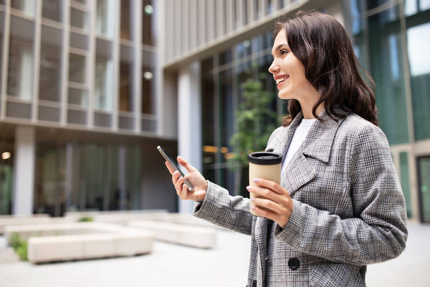 Бізнес-додаток, комунікаційна пропозиція. Молода бізнес-леді тримає мобільний телефон і чашку кави, посміхаючись стоїть в міському районі Поруч Бізнес-центр Будівництво зовні, вид збоку - Фото, зображення
