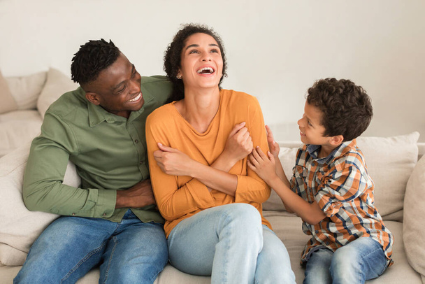 Diversa familia divirtiéndose juntos, papá e hijo cosquillas mamá riéndose sentado en el sofá en casa. Afro-americano papá, árabe mamá y su pequeño niño disfrutando de jugar y acurrucarse - Foto, imagen