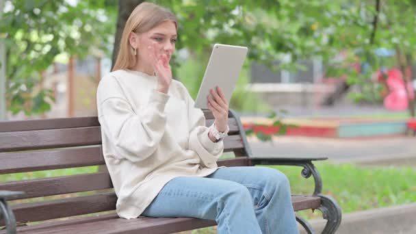Mujer joven haciendo video chat en la tableta mientras está sentada al aire libre en un banco - Imágenes, Vídeo