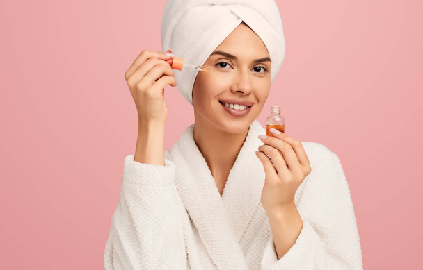 Happy young female in bathrobe and towel with towel on head applying moisturizing hyaluronic acid as skincare procedure on pink background - Φωτογραφία, εικόνα