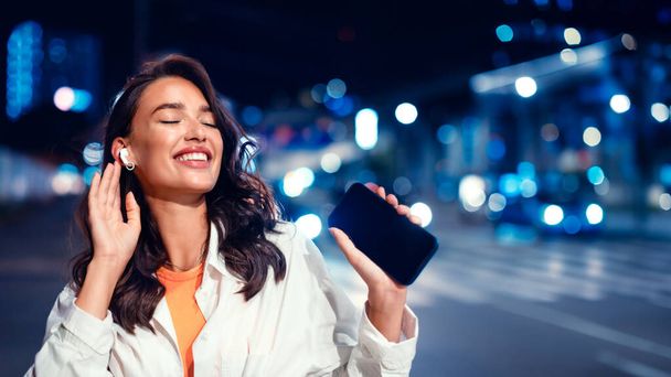 Радостная дама, наслаждающаяся любимой музыкой в наушниках со смартфоном, гуляющая ночью по городской улице с неоновой подсветкой на заднем плане, панорама с бесплатным пространством - Фото, изображение