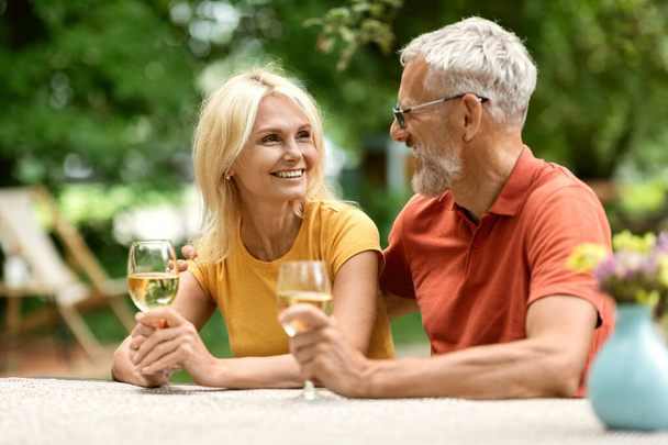 Portret van liefdevol getrouwd senior paar drinken wijn buiten, gelukkig volwassen echtgenoten zitten aan tafel in de tuin, picknick buiten, kijken en glimlachen naar elkaar, vieren verjaardag - Foto, afbeelding