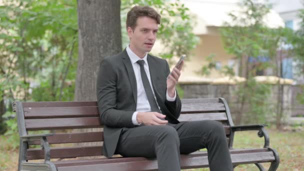 Nuori liikemies puhuu puhelimessa istuessaan ulkona penkillä - Materiaali, video