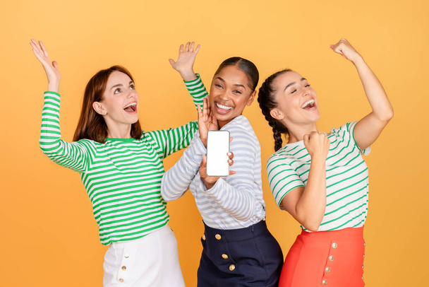 Zorgeloos aantrekkelijke millennial dames in stijlvolle casual outfits hebben plezier samen op oranje studio achtergrond, met smartphone met leeg scherm, dansen en lachen, mockup, panorama. E-handel - Foto, afbeelding