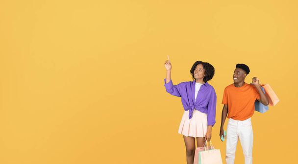 Bin yıllık siyahi kadın ve erkek paketleri boş alanı işaret ediyor, alışveriş yapmayı öneriyor, sarı stüdyo arka planında izole edilmiş. Reklam ve teklif, satış, bir fikrim var - Fotoğraf, Görsel