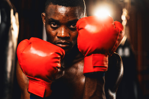 Boxkämpfer in hemdsloser Pose, afroamerikanischer schwarzer Boxer mit rotem Handschuh in defensiver Wachhaltung, bereit zum Kampf und zum Schlagabtausch im Fitnessstudio mit Kickbag und Boxausrüstung im Hintergrund. Impulse - Foto, Bild