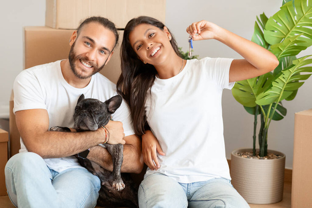 Positive happy loving young multicultural couple enjoying their new apartment, cheerful man and woman bought house, sit on floor by carton boxes, hugging their dog, showing key. Relocation concept - Photo, Image