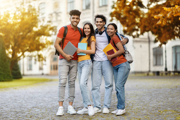 Grupo de amigos multiétnicos de la universidad con libros de trabajo posando al aire libre en el campus, alegres jóvenes estudiantes felices abrazando y mirando a la cámara, divirtiéndose mientras camina después de clases, copiar el espacio - Foto, Imagen