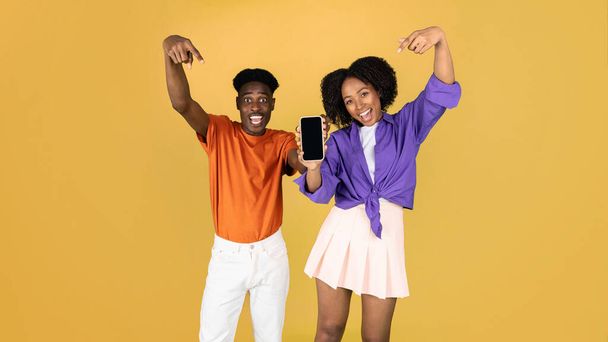 Glad excited millennial black guy and lady point fingers at smartphone with blank screen, recommend app, isolated on yellow studio background, panorama. Gadget and app, website, blog, ad and offer - Photo, Image