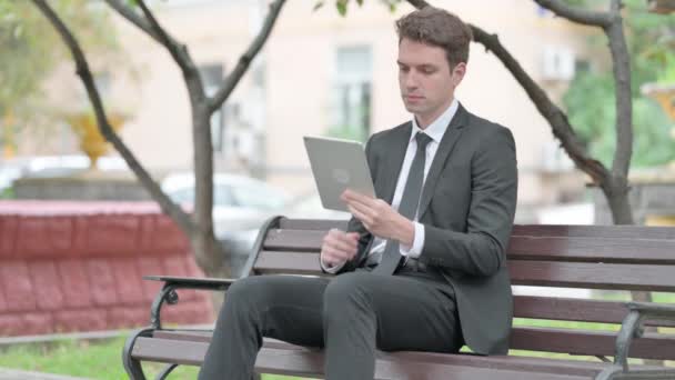 Бизнесмен, сидя на скамейке, пользуется планшетом - Кадры, видео