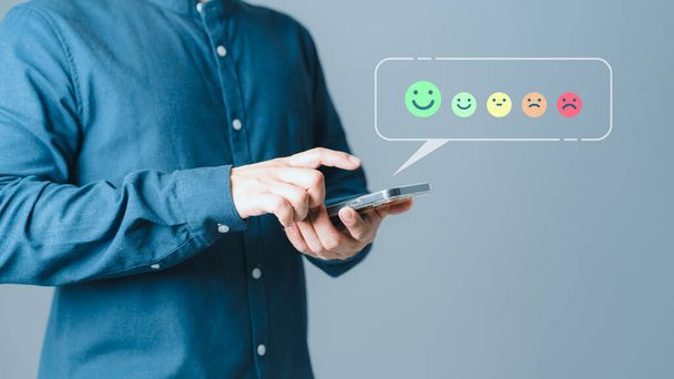 Ügyfélkezek segítségével egy mobiltelefon felugró mosoly arc ikon visszajelzést felülvizsgálat elégedettségi szolgáltatás véleménye és ajánlólevél. Ügyfélszolgálati tapasztalat felmérése és üzleti elégedettség felmérése. - Fotó, kép