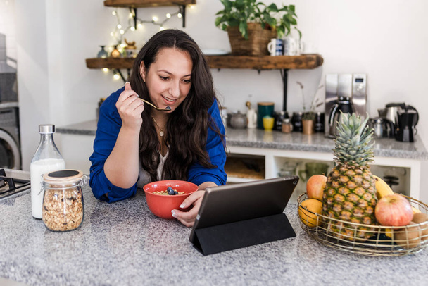 jovem mulher latina com sobrepeso usando tablet enquanto toma café da manhã em casa cozinha no México América Latina, hispânico plus size female - Foto, Imagem