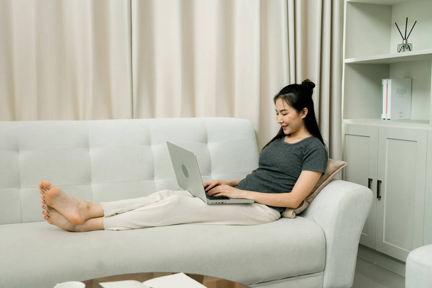 Asyalı kadın evde çalışmak için dizüstü bilgisayar kullanarak kanepeye oturuyor.. - Fotoğraf, Görsel
