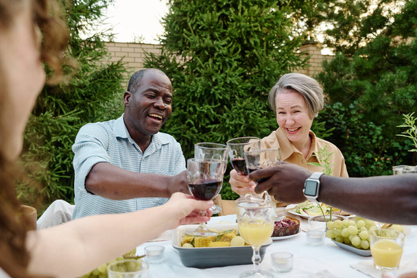 Gelukkig volwassen intercultureel paar met glazen zelfgemaakte rode wijn toasten met hun zoon en zijn vrouw over tafel geserveerd voor het diner - Foto, afbeelding