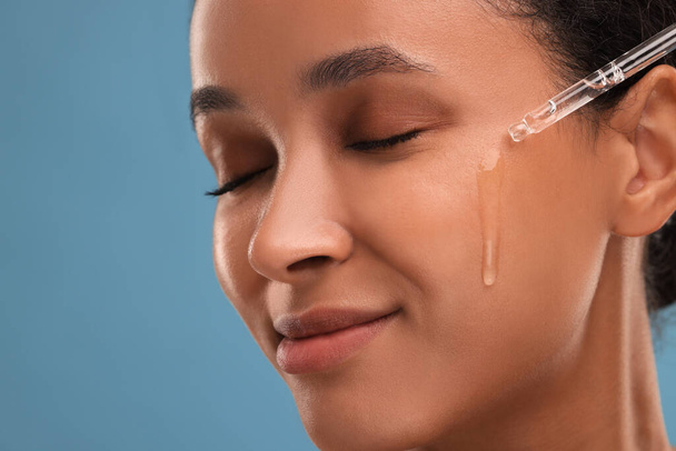 Beautiful woman applying serum onto her face on blue background, closeup - Foto, Imagem