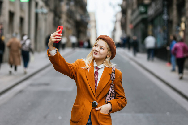 Mujer asiática con estilo tomando selfies con teléfono móvil de pie en la calle. Tecnología y concepto de redes sociales. - Foto, Imagen